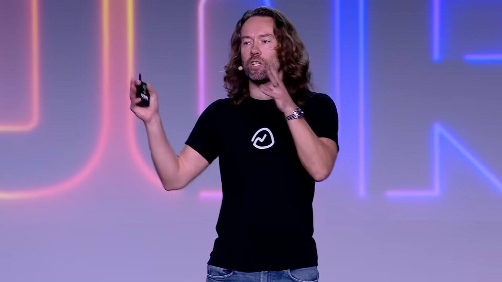 DHH on stage at Rails World 2026 in Austin, wearing a Basecamp t-shirt and holding a presentation clicker in front of a neon-lit backdrop