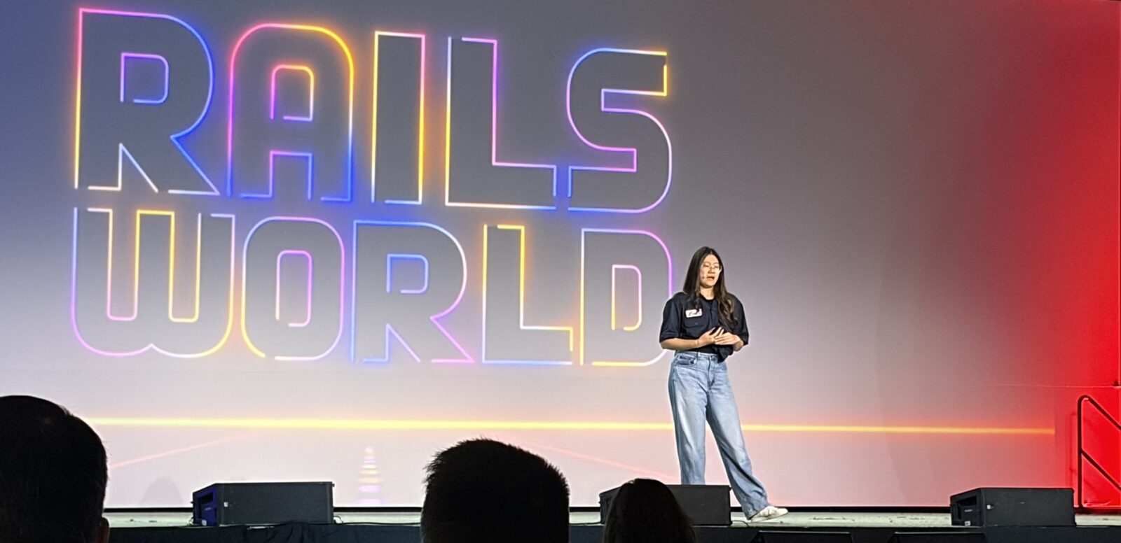 Jenny Shen presenting on stage at Rails World 2026, in front of a large screen showing the glowing 'Rails World' logo